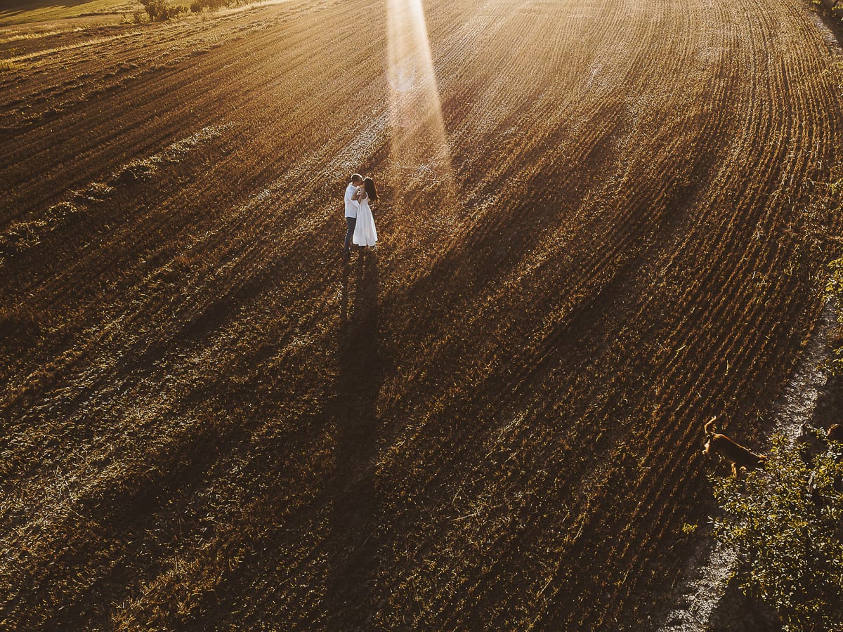 Prewedding romantico e da sogno al tramonto in Monferrato. Valeria e Omar camminano tenendosi per mano in un campo di grano tagliato. Foto aerea ripresa dal drone.