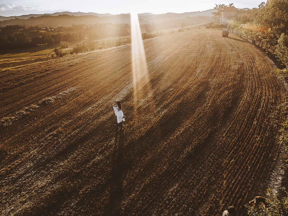 Prewedding romantico e da sogno al tramonto in Monferrato. Valeria e Omar camminano tenendosi per mano in un campo di grano tagliato. Foto aerea ripresa dal drone.
