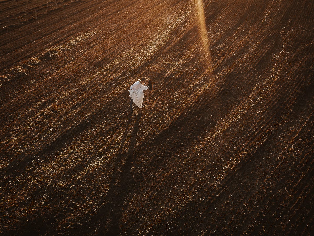 Prewedding romantico e da sogno al tramonto in Monferrato. Valeria e Omar camminano tenendosi per mano in un campo di grano tagliato. Foto aerea ripresa dal drone.