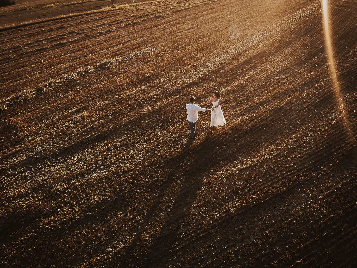 Prewedding romantico e da sogno al tramonto in Monferrato. Valeria e Omar camminano tenendosi per mano in un campo di grano tagliato. Foto aerea ripresa dal drone.
