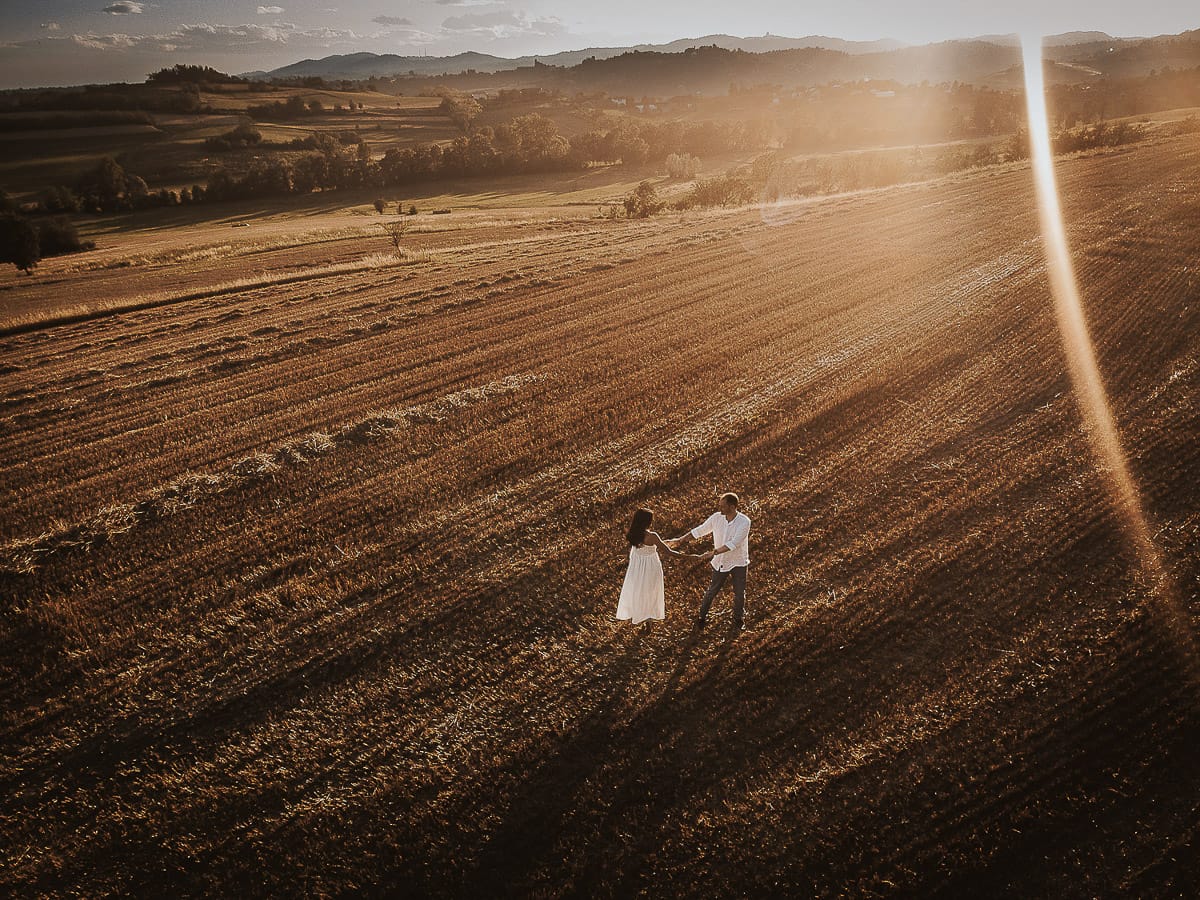 Prewedding romantico e da sogno al tramonto in Monferrato. Valeria e Omar camminano tenendosi per mano in un campo di grano tagliato. Foto aerea ripresa dal drone.