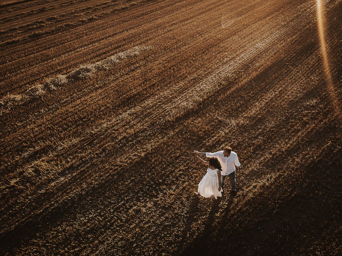 Prewedding romantico e da sogno al tramonto in Monferrato. Valeria e Omar camminano tenendosi per mano in un campo di grano tagliato. Foto aerea ripresa dal drone.