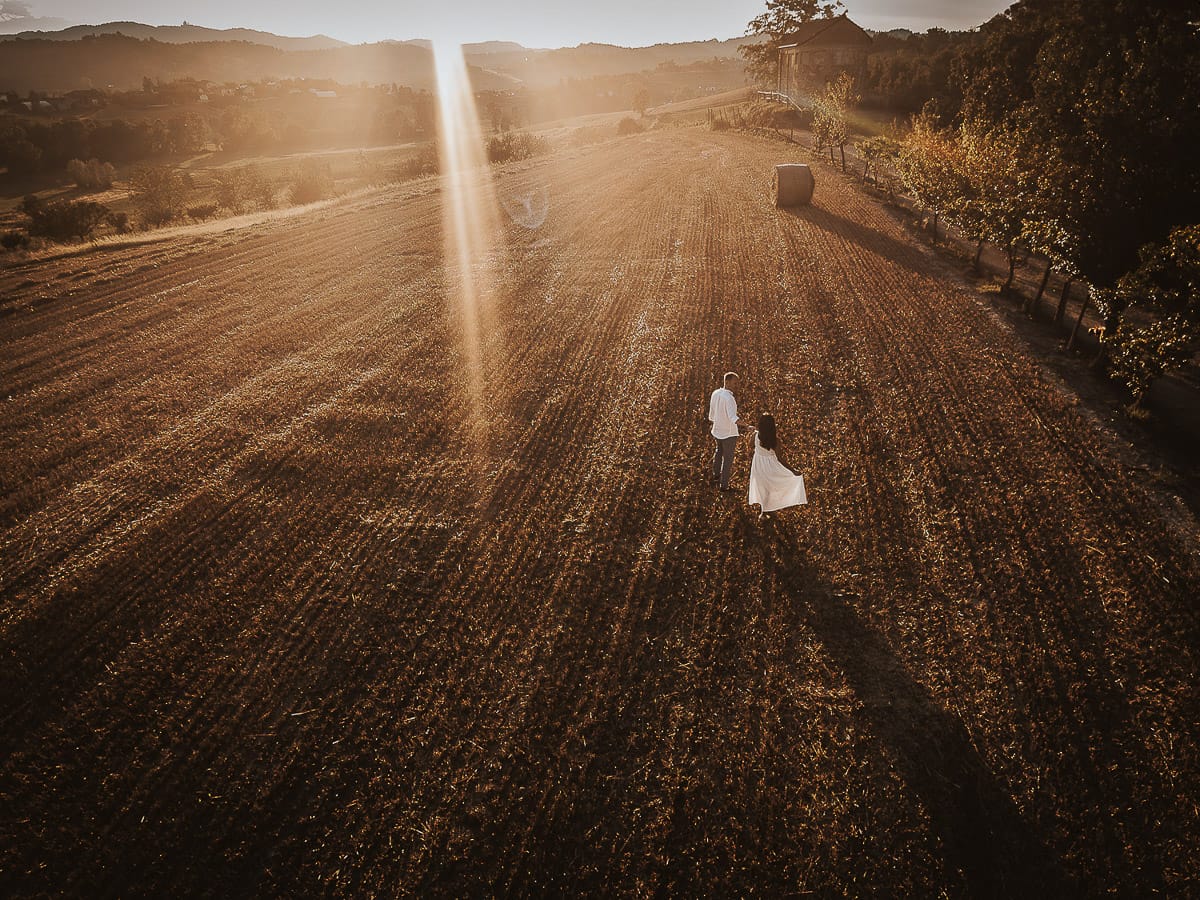 Prewedding romantico e da sogno al tramonto in Monferrato. Valeria e Omar camminano tenendosi per mano in un campo di grano tagliato. Foto aerea ripresa dal drone.