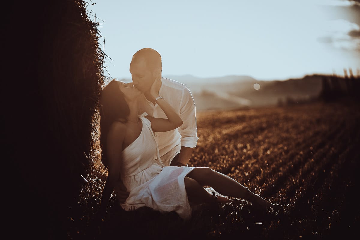 Prewedding romantico e da sogno al tramonto in Monferrato. Valeria e Omar seduti in un campo di grano tagliato vicino ad una rotoballa si baciano in controluce
