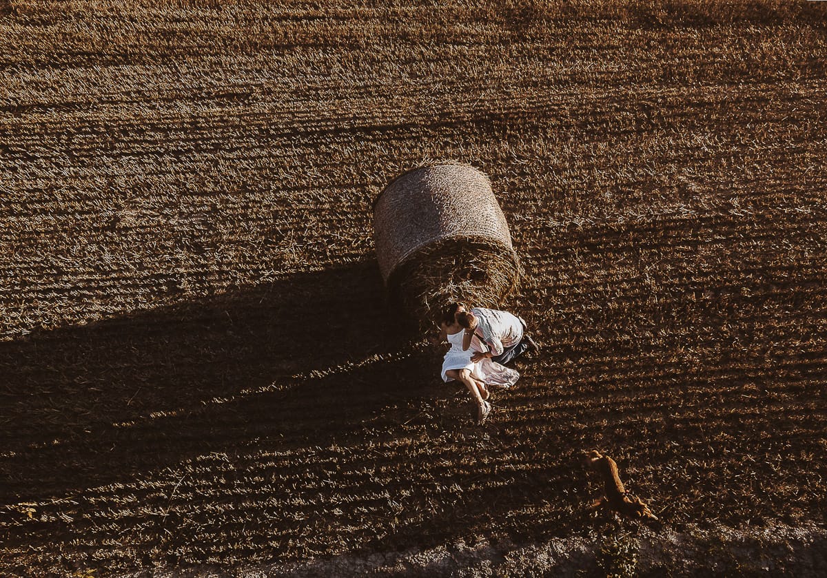 Prewedding romantico e da sogno al tramonto in Monferrato. Valeria e Omar seduti in un campo di grano tagliato vicino ad una rotoballa si baciano in controluce con la cagnolina vicino a loro. Foto aerea ripresa dal drone.