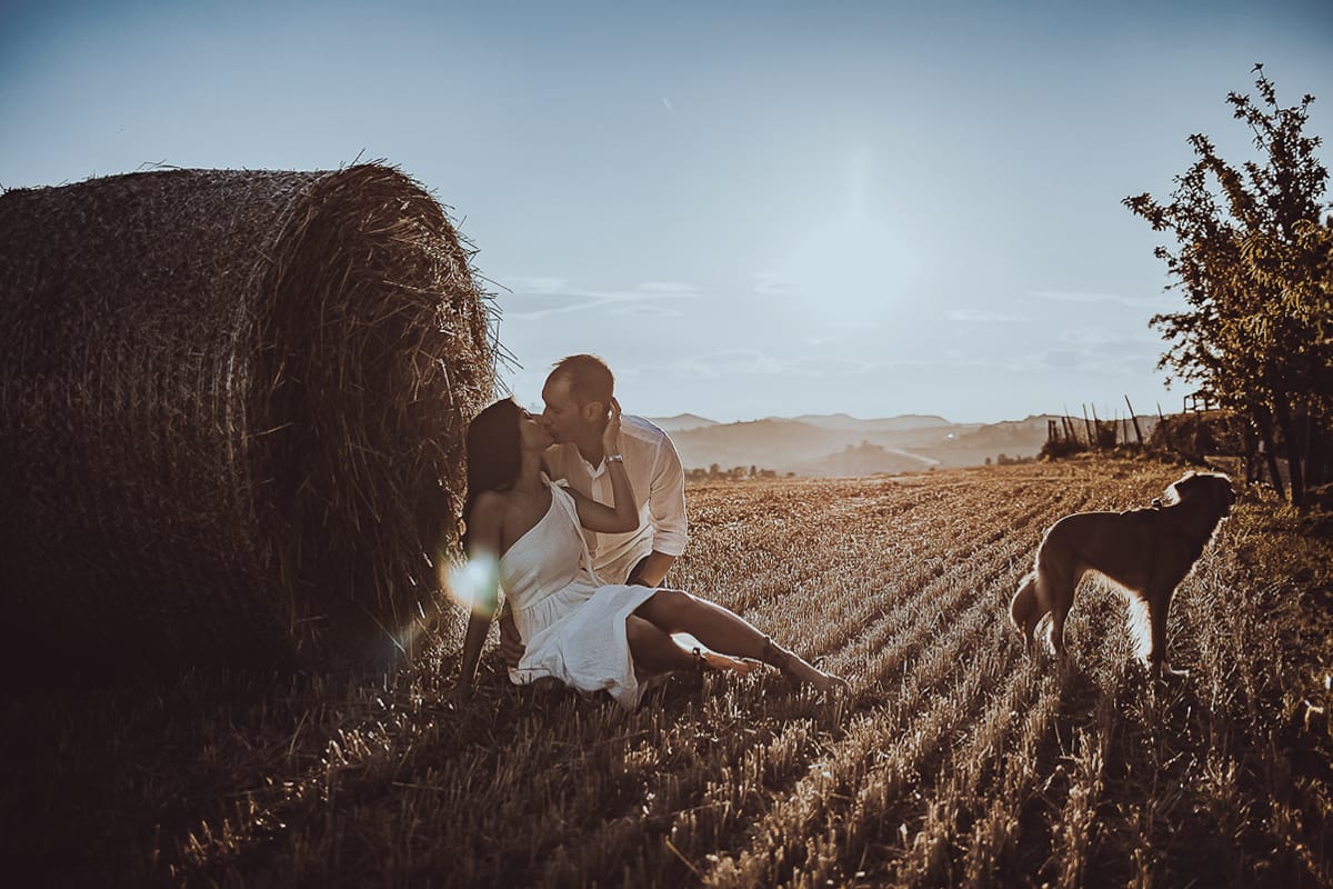 Prewedding romantico e da sogno al tramonto in Monferrato. Valeria e Omar seduti in un campo di grano tagliato vicino ad una rotoballa si baciano in controluce con la cagnolina vicino a loro