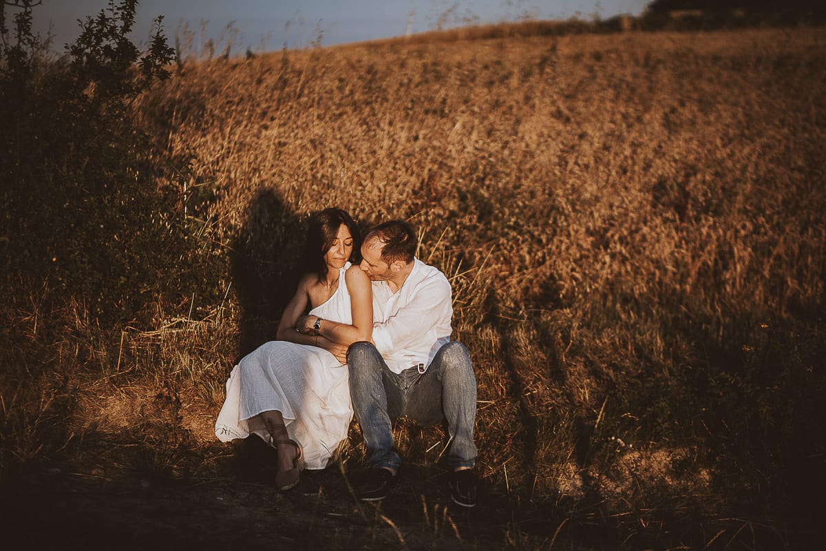 Prewedding romantico e da sogno al tramonto in Monferrato. Valeria e Omar seduti al bordo di un campo di grano. Omar bacia Valeria su di Una spalla