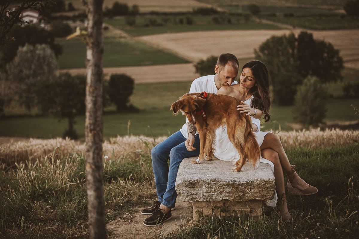 Prewedding romantico e da sogno al tramonto in Monferrato. Valeria e Omar seduti su di una panchina di pietra. Omar bacia la spalla di Valeria. La cagnolina Nadine è con loro.