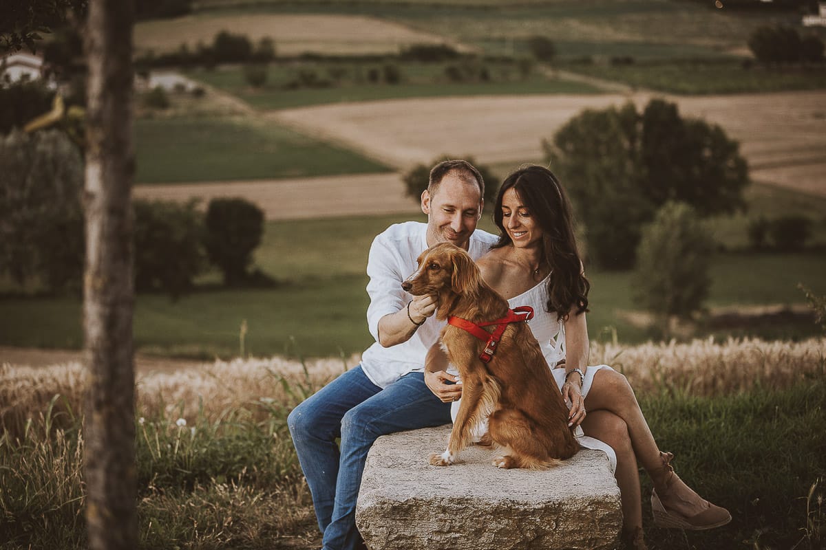 Prewedding romantico e da sogno al tramonto in Monferrato. Valeria e Omar seduti su di una panchina di pietra. Omar bacia la spalla di Valeria. la cagnolina Nadine è davanti a loro.
