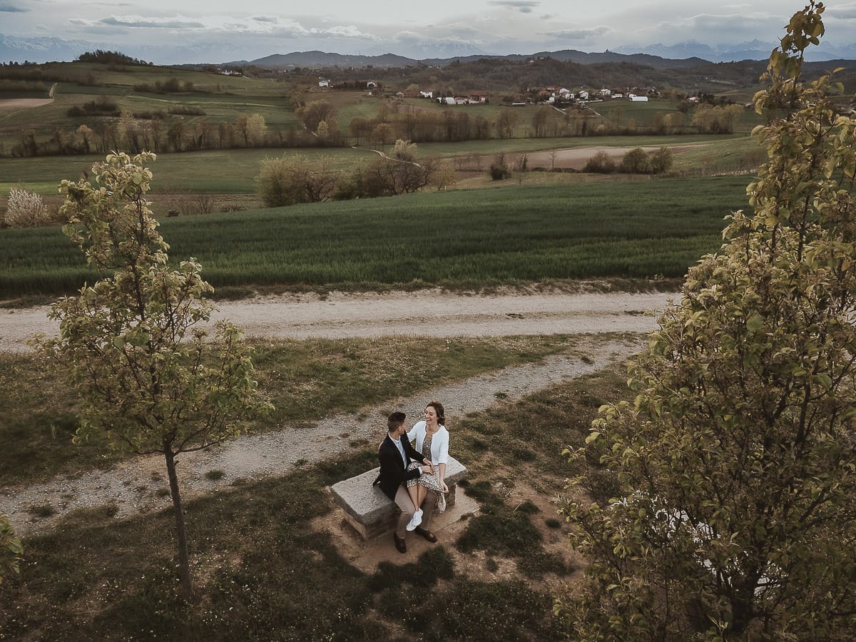 Prewedding primaverile in collina a Mombello di Torino di Federica e Mattia. La coppia è seduta sulla panchina di pietra e si guarda. Foto ripresa dal drone.