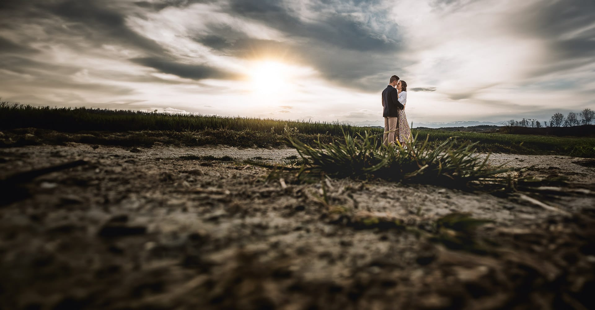 Prewedding primaverile a Mombello di Torino di Federica e Mattia. La coppia abbracciata è ripresa dal drone posto sul terreno dietro un ciuffo d'erba. Ci sono grandi nuvole e una bellissima luce.
