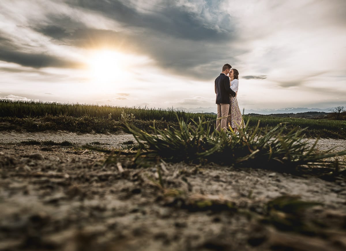 Prewedding primaverile in collina a Mombello di Torino di Federica e Mattia. Ritratto controluce con il sole tra di loro che sta tramontando fra le nuvole- foto ripresa dal drone posato sul suolo dietro un ciuffo d'erba.