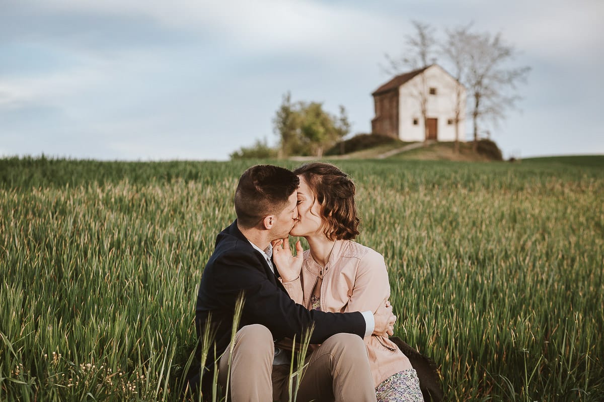 Prewedding primaverile in collina a Mombello di Torino di Federica e Mattia. Federica e Mattia sono seduti in un campo di grano ancora verde. Dietro la bellissima collina con la chiesetta di San Lorenzo.