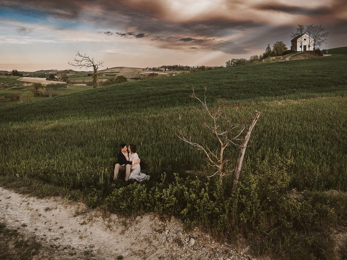 Prewedding primaverile in collina a Mombello di Torino di Federica e Mattia. Federica e Mattia sono seduti in un campo di grano ancora verde. Dietro la bellissima collina con la chiesetta di San Lorenzo. Foto dal drone.