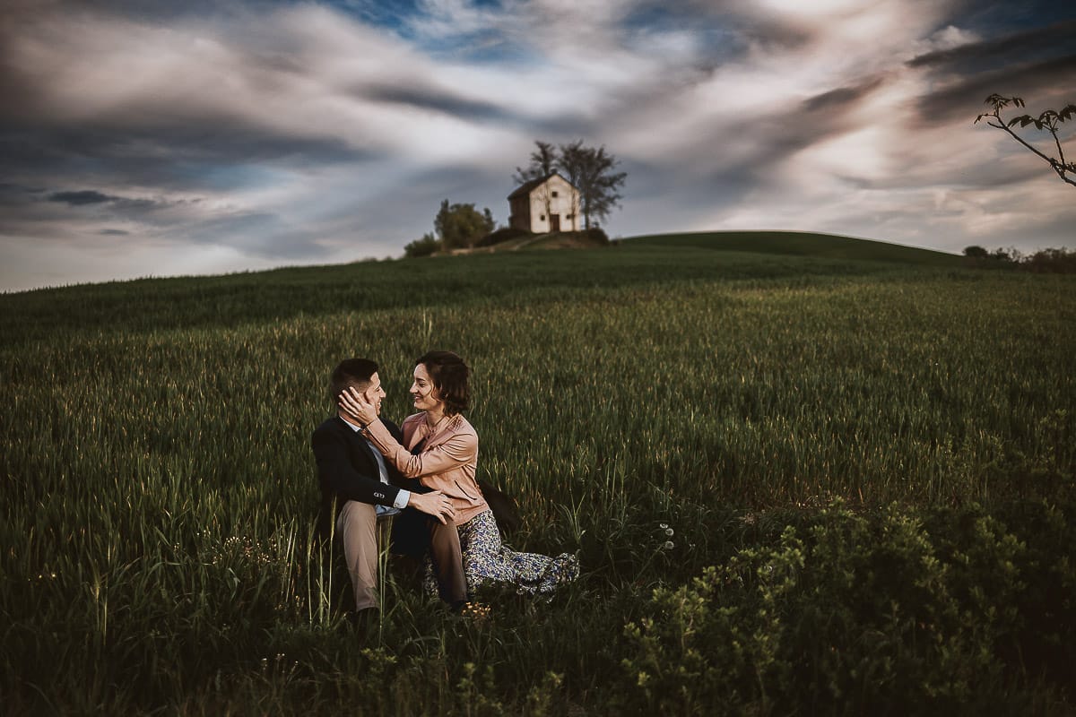 Prewedding primaverile in collina a Mombello di Torino di Federica e Mattia. Federica e Mattia sono seduti in un campo di grano ancora verde. Dietro la bellissima collina con la chiesetta di San Lorenzo.