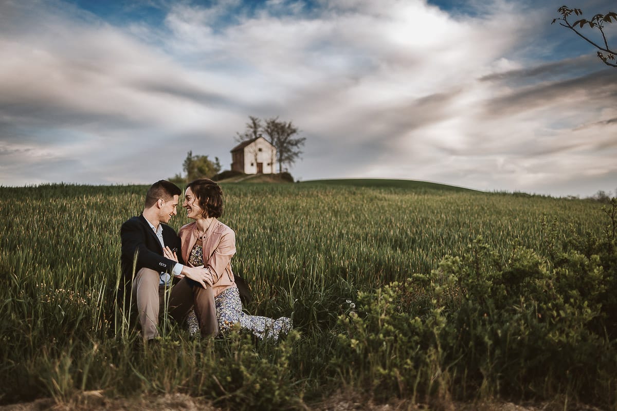 Prewedding primaverile in collina a Mombello di Torino di Federica e Mattia. Federica e Mattia sono seduti in un campo di grano ancora verde. Dietro la bellissima collina con la chiesetta di San Lorenzo.