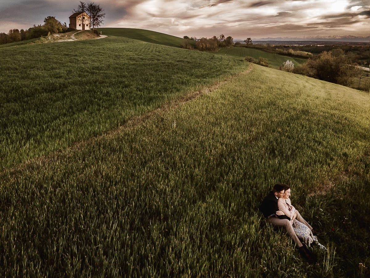 Prewedding primaverile in collina a Mombello di Torino di Federica e Mattia. Federica e Mattia sono seduti in un campo di grano ancora verde. Dietro la bellissima collina con la chiesetta di San Lorenzo. Foto dal drone.