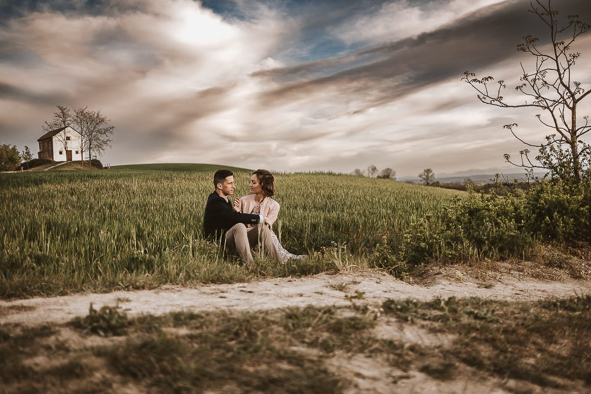 Prewedding primaverile in collina a Mombello di Torino di Federica e Mattia. Federica e Mattia sono seduti in un campo di grano ancora verde. Dietro la bellissima collina con la chiesetta di San Lorenzo.