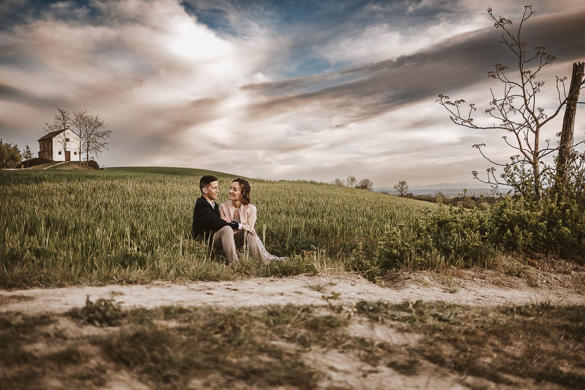 Prewedding primaverile in collina a Mombello di Torino di Federica e Mattia. Federica e Mattia sono seduti in un campo di grano ancora verde. Dietro la bellissima collina con la chiesetta di San Lorenzo.