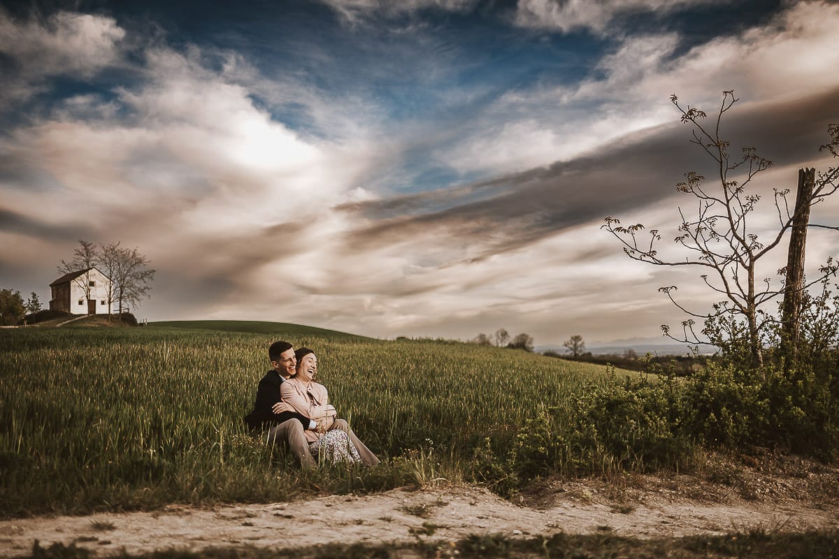 Prewedding primaverile in collina a Mombello di Torino di Federica e Mattia. Federica e Mattia sono seduti in un campo di grano ancora verde. Dietro la bellissima collina con la chiesetta di San Lorenzo.