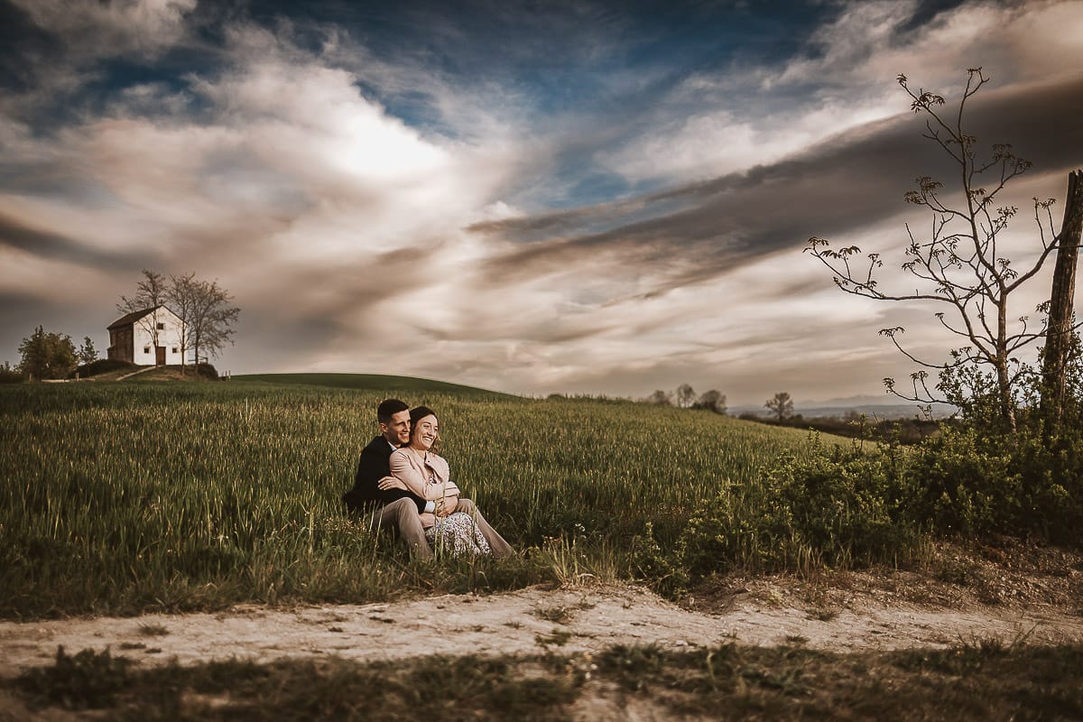 Prewedding primaverile in collina a Mombello di Torino di Federica e Mattia. Federica e Mattia sono seduti in un campo di grano ancora verde. Dietro la bellissima collina con la chiesetta di San Lorenzo.
