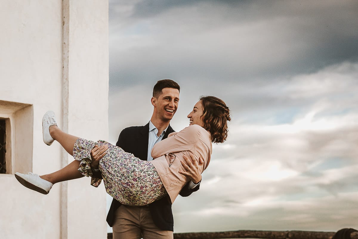 Prewedding primaverile in collina a Mombello di Torino di Federica e Mattia. Federica e Mattia di fronte alla chiesa di San Lorenzo. Mattia prende in braccio Federica.