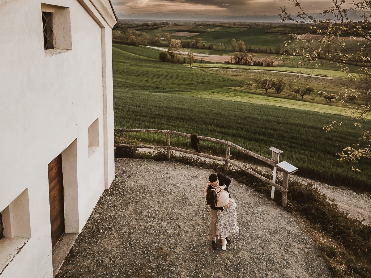 Prewedding primaverile in collina a Mombello di Torino di Federica e Mattia. Federica e Mattia di fronte alla chiesa di San Lorenzo. Mattia prende in braccio Federica. Foto dal drone.