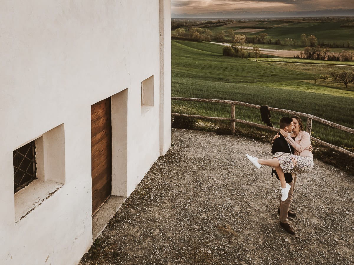 Prewedding primaverile in collina a Mombello di Torino di Federica e Mattia. Federica e Mattia di fronte alla chiesa di San Lorenzo. Mattia prende in braccio Federica. Foto dal drone