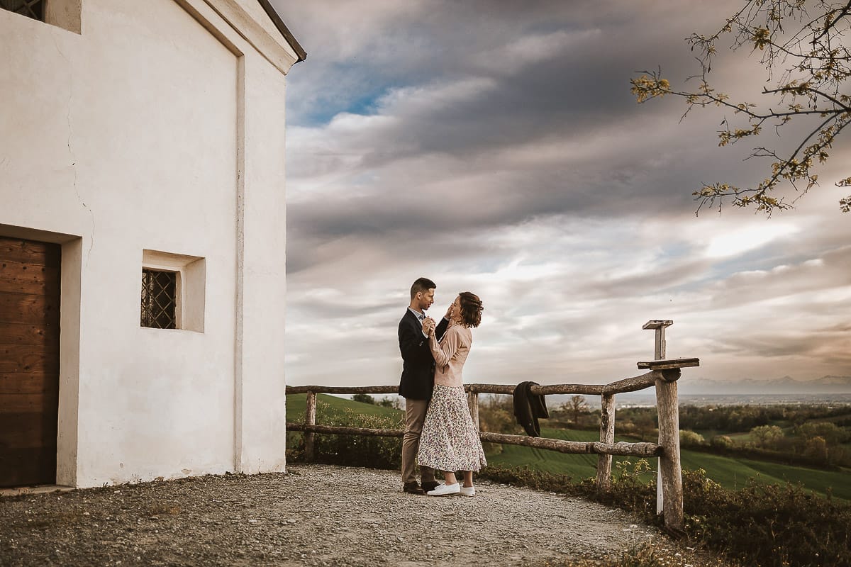 Prewedding primaverile in collina a Mombello di Torino di Federica e Mattia. Federica e Mattia di fronte alla chiesa di San Lorenzo. Mattia e Federica si abbracciano.