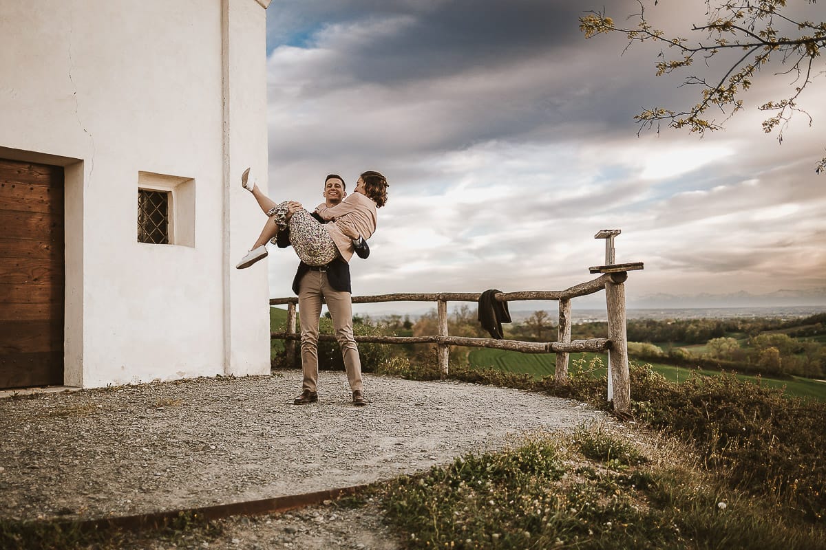 Prewedding primaverile in collina a Mombello di Torino di Federica e Mattia. Federica e Mattia di fronte alla chiesa di San Lorenzo. Mattia prende in braccio Federica.