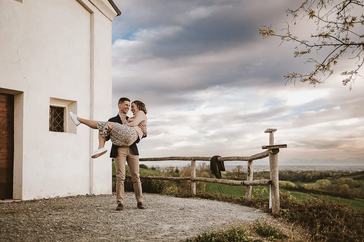 Prewedding primaverile in collina a Mombello di Torino di Federica e Mattia. Federica e Mattia di fronte alla chiesa di San Lorenzo. Mattia prende in braccio Federica.