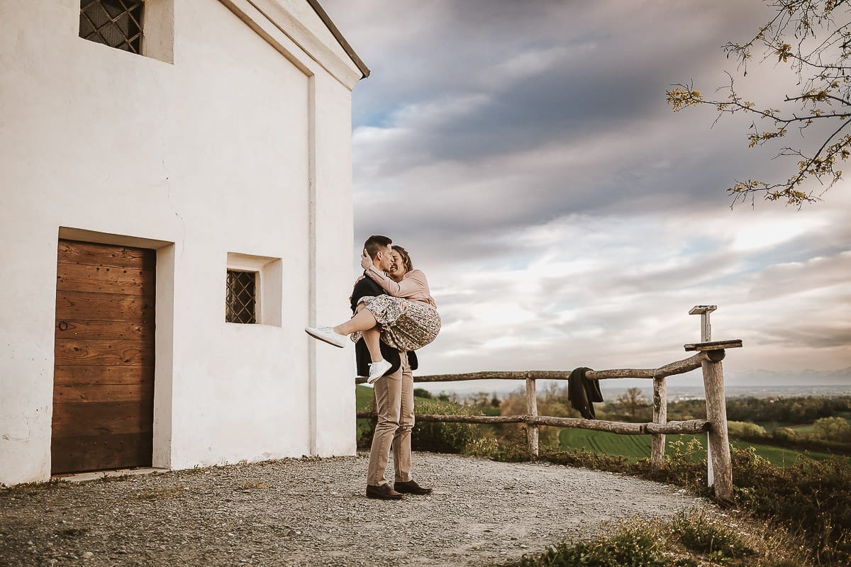Prewedding primaverile in collina a Mombello di Torino di Federica e Mattia. Federica e Mattia di fronte alla chiesa di San Lorenzo. Mattia prende in braccio Federica.