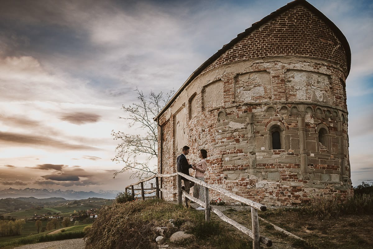 Prewedding primaverile in collina a Mombello di Torino di Federica e Mattia. Federica e Mattia di fronte alla chiesa di San Lorenzo. Mattia e Federica appoggiati alla staccionata con la chiesa romanica sullo sfondo.