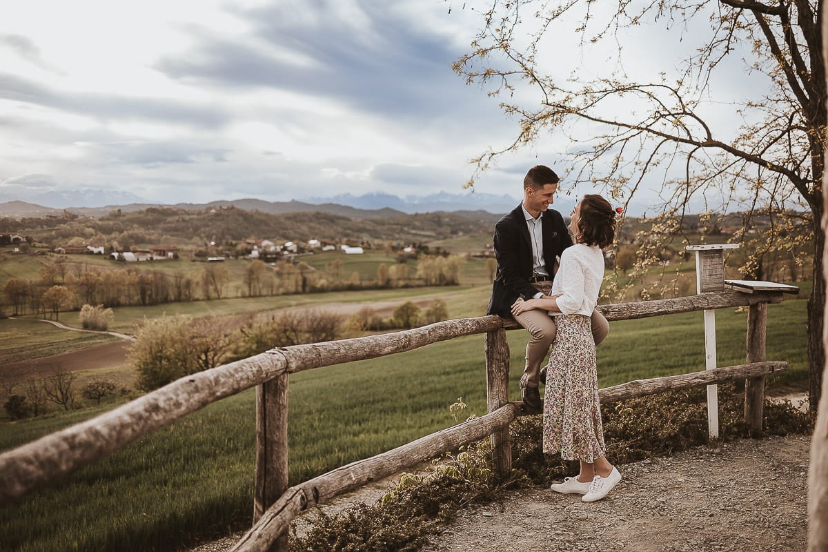 Prewedding primaverile in collina a Mombello di Torino di Federica e Mattia. Mattia è seduto sulla staccionata di legno e Federica lo guarda.