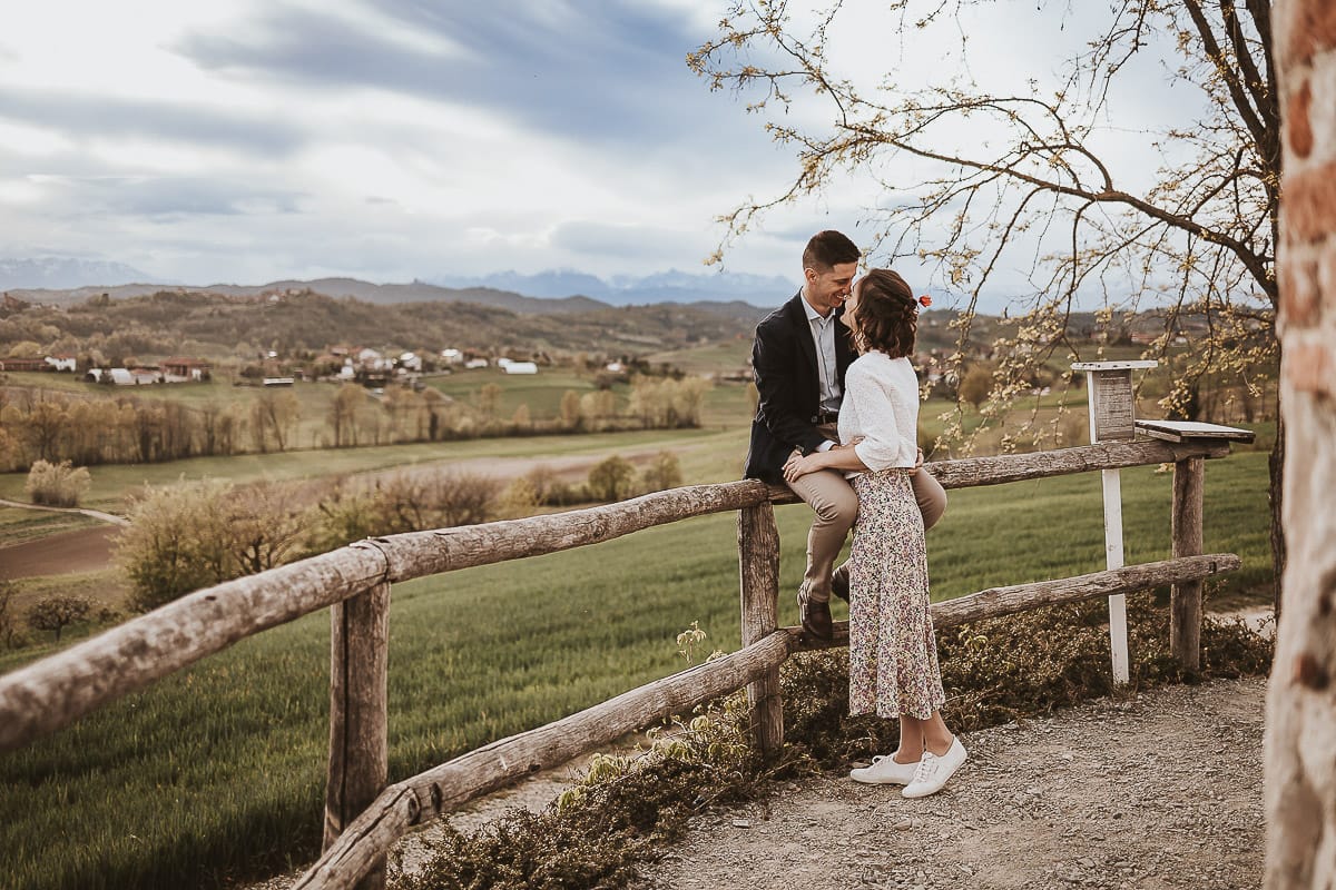 Prewedding primaverile in collina a Mombello di Torino di Federica e Mattia. Mattia è seduto sulla staccionata e Federica lo bacia.