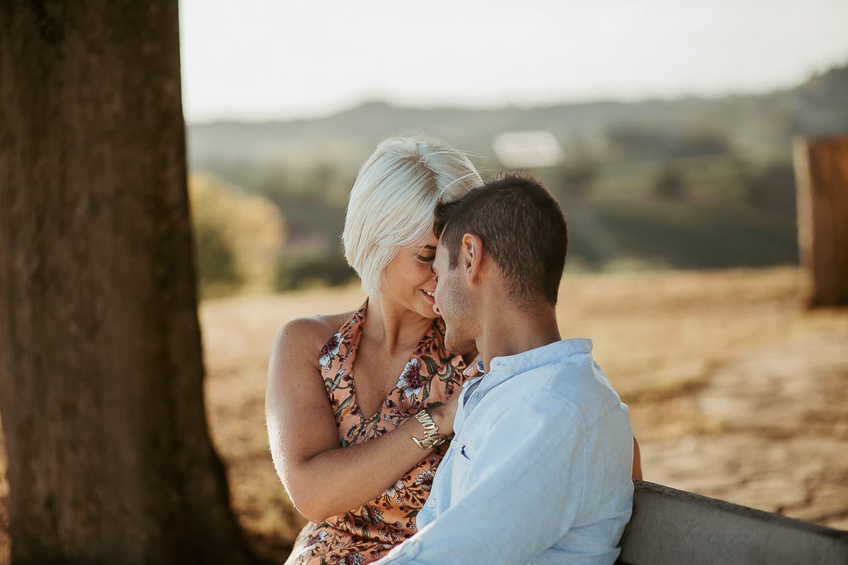 Prewedding da sogno a Nizza Monferrato intorno alla chiesa della Madonna della Neve. Matteo e Fiammetta seduti su di una panchina all'ombra di un grande albero., si guardano intensamente