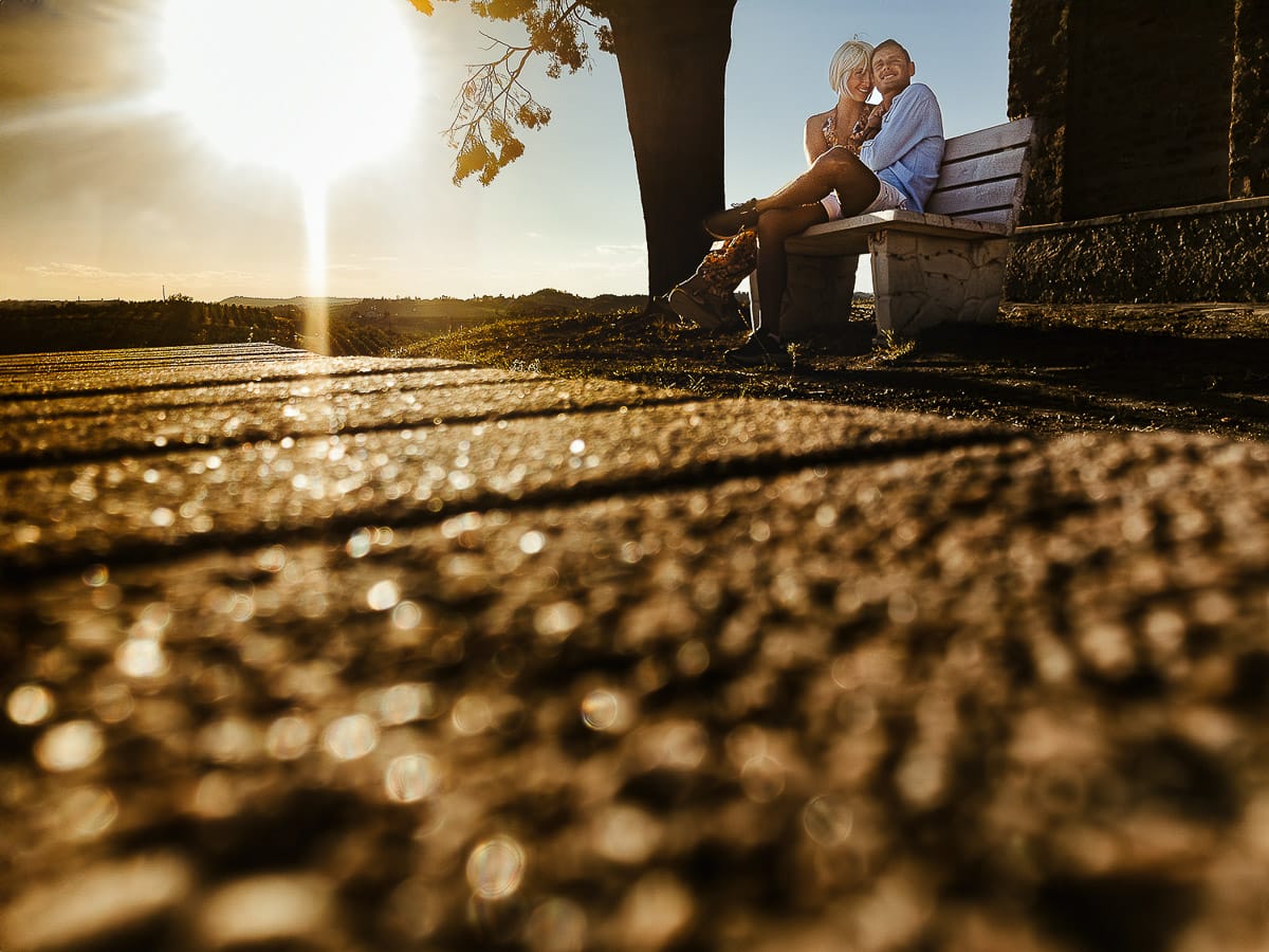 Prewedding da sogno a Nizza Monferrato intorno alla chiesa della Madonna della Neve. Matteo e Fiammetta seduti su di una panchina all'ombra di un grande albero. Foto in controluce ripresa dal drone posto sul terreno.