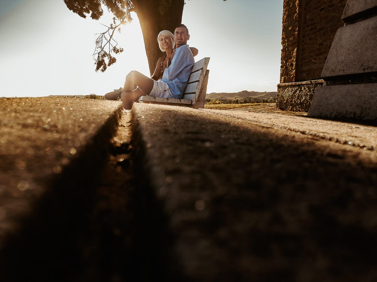 Prewedding da sogno a Nizza Monferrato intorno alla chiesa della Madonna della Neve. Matteo e Fiammetta seduti su di una panchina all'ombra di un grande albero. Foto in controluce ripresa dal drone posto sul terreno.