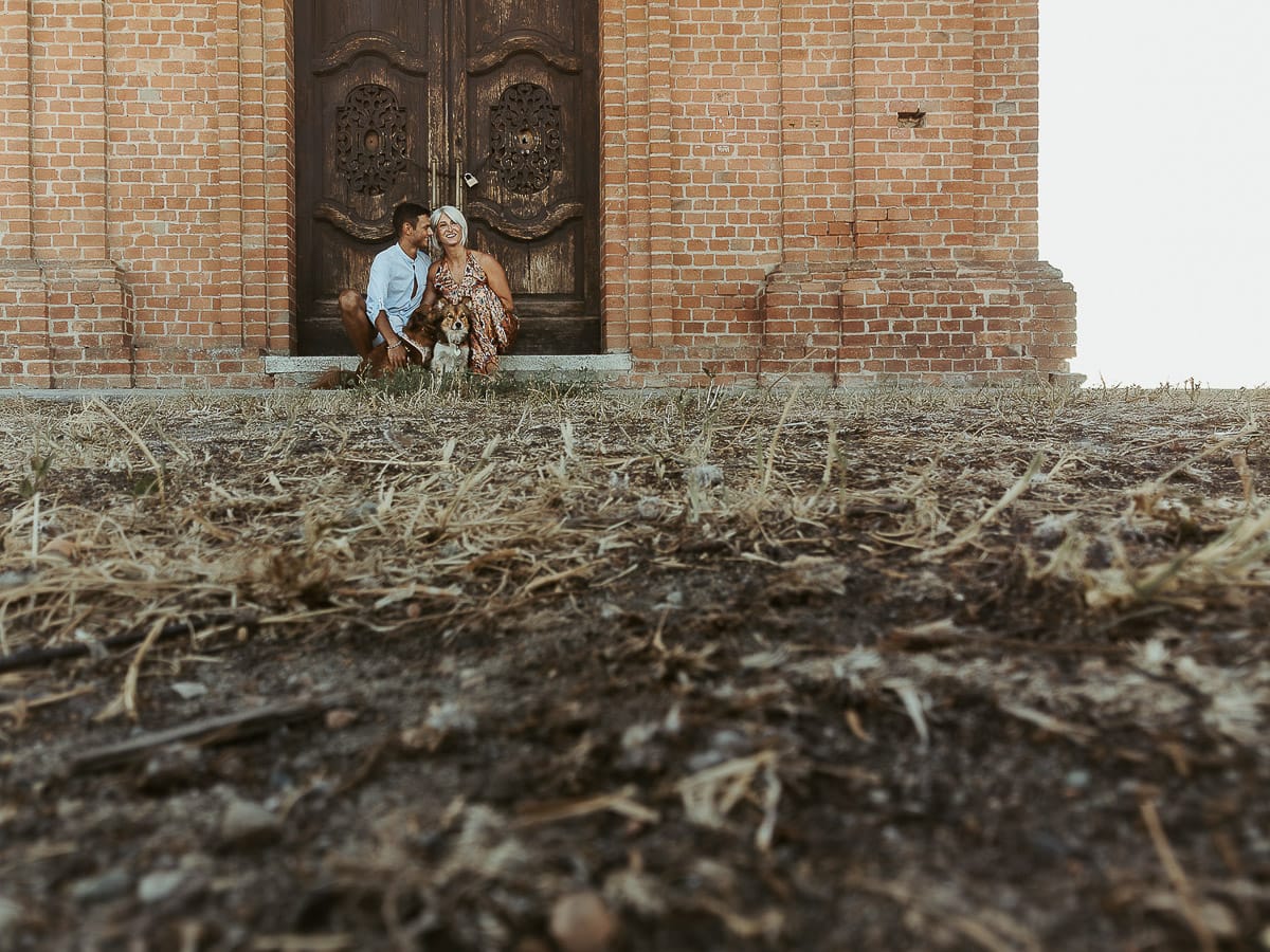 Prewedding da sogno a Nizza Monferrato intorno alla chiesa della Madonna della Neve. Fiammetta e Matteo seduti sul gradino del portone della chiesa con i loro volpini Megan e Harry Foto scattata dal drone posto sul terreno.