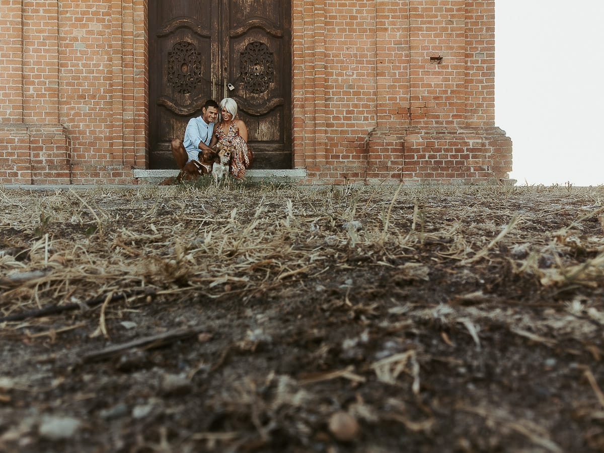 Prewedding da sogno a Nizza Monferrato intorno alla chiesa della Madonna della Neve. Fiammetta e Matteo seduti sul gradino del portone della chiesa con i loro volpini Megan e Harry Foto scattata dal drone posto sul terreno.