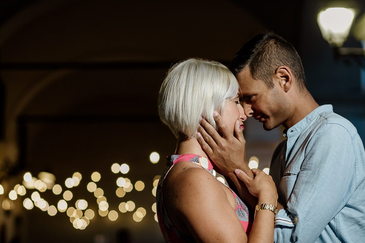 Prewedding da sogno a Nizza Monferrato nel centro storico. Fiammetta e Matteo davanti al bellissimo portone scolpito di un palazzo. storico. Flash off camera usato in modo sapiente illumina la coppia quando è ormai sera. Dietro di loro fili di luci che creano bokeh.