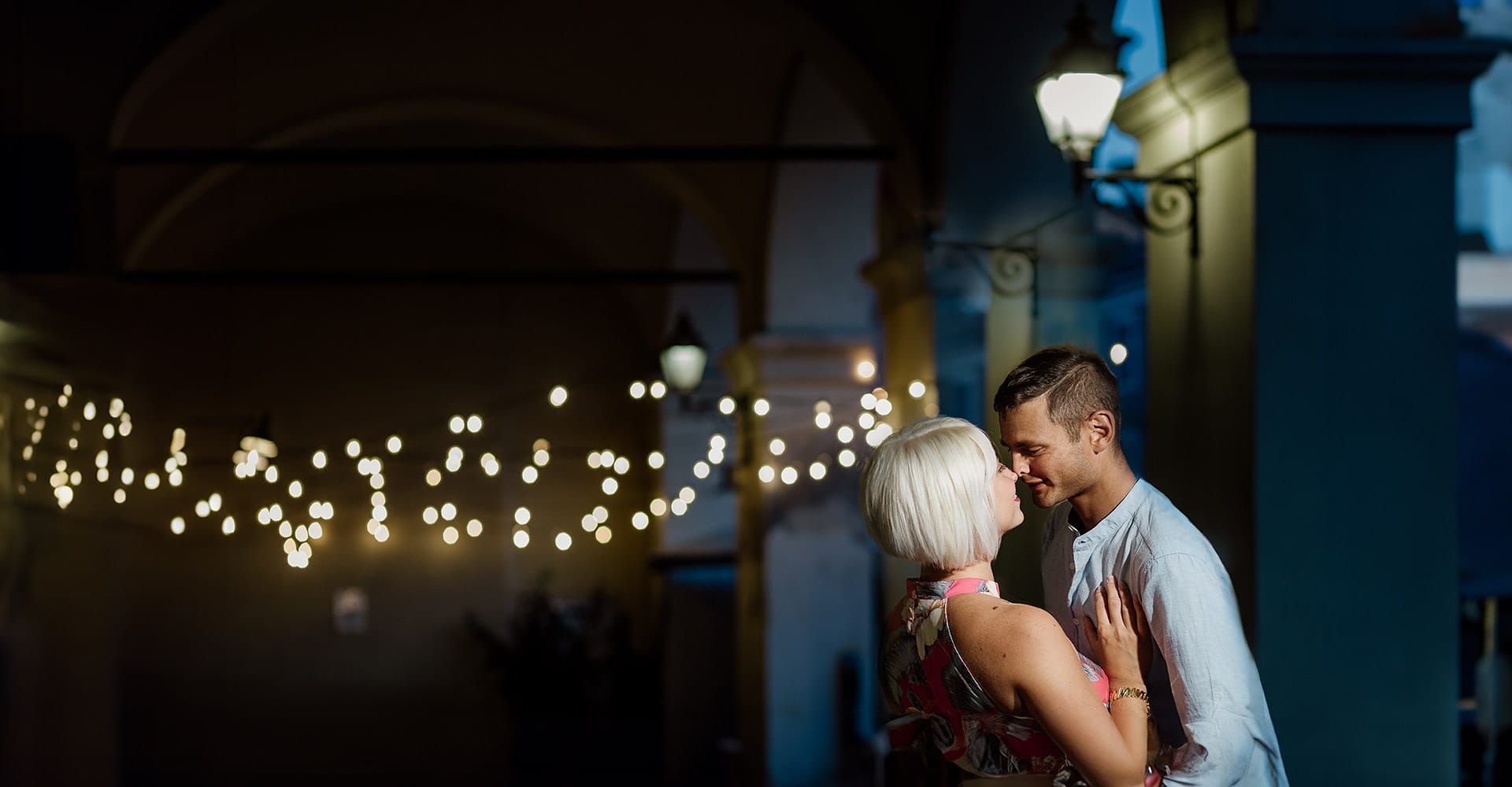 Prewedding da sogno a Nizza Monferrato nel centro storico. Fiammetta e Matteo davanti al bellissimo portone scolpito di un palazzo. storico. Flash off camera usato in modo sapiente illumina la coppia quando è ormai sera. Dietro di loro fili di luci che creano bokeh.