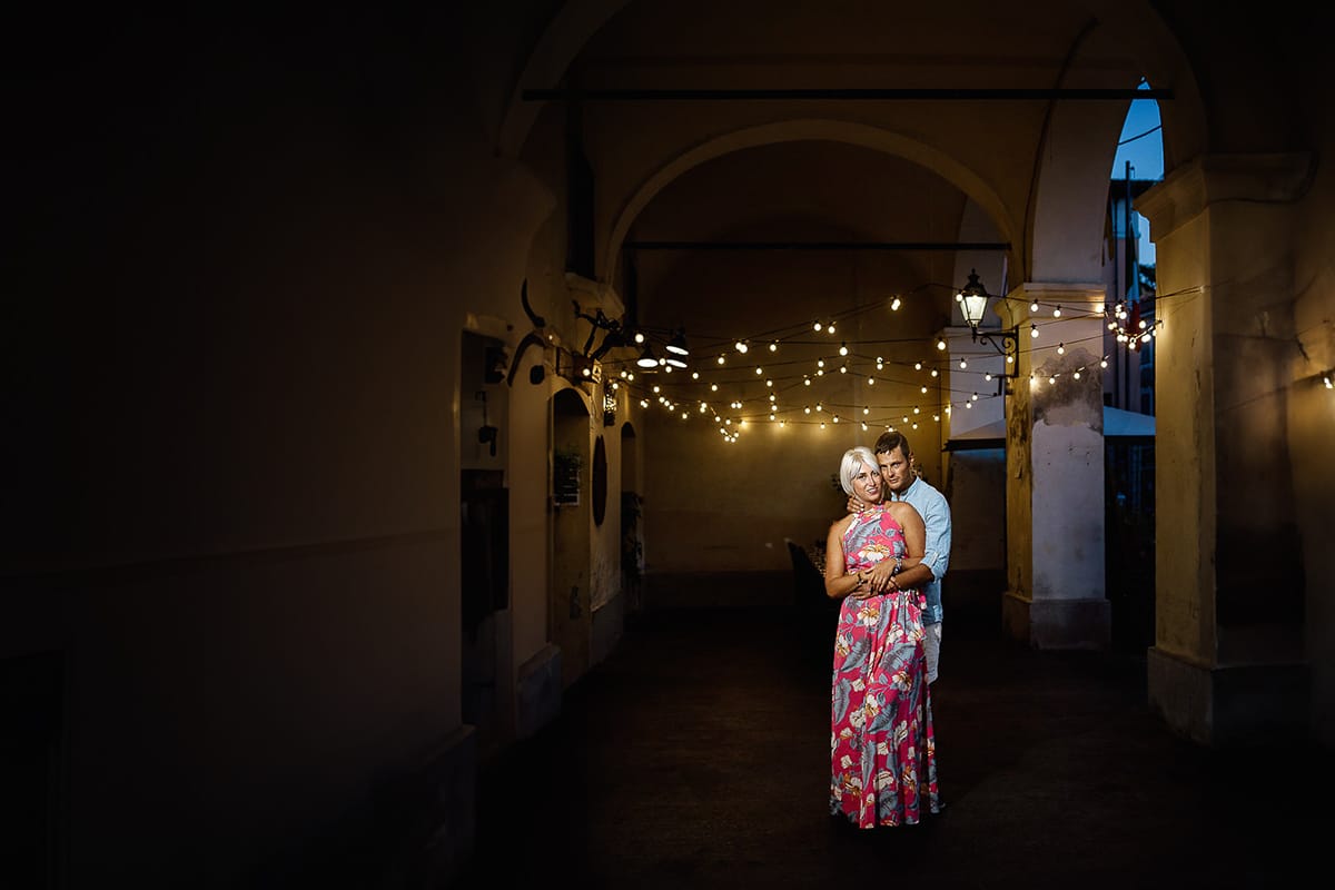 Prewedding da sogno a Nizza Monferrato nel centro storico. Fiammetta e Matteo davanti al bellissimo portone scolpito di un palazzo. storico. Flash off camera usato in modo sapiente illumina la coppia quando è ormai sera. Dietro di loro fili di luci che creano bokeh.