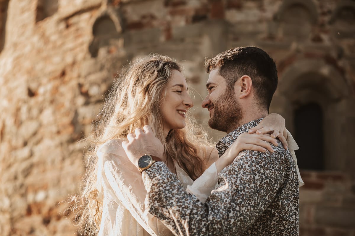 Anita e Davide, si accarezzano per mano davanti a una chiesetta romanica a Mombello di Torino, immersi nella calda luce del sole primaverile del Monferrato
