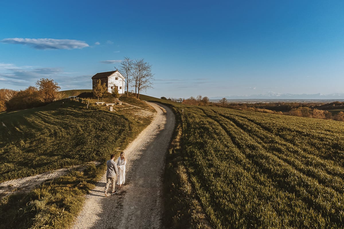Un drone riprende la suggestiva prospettiva dall'alto di Anita e Davide, futuri sposi, che passeggiano felici davanti alla chiesetta romanica di San Lorenzo a Mombello di Torino. I colori vivaci della primavera e le colline del Monferrato fanno da sfondo a questo momento indimenticabile.