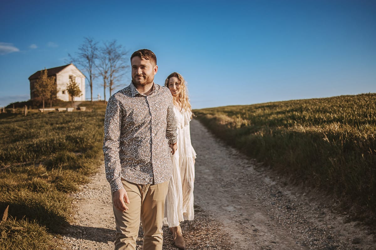 Anita e Davide, futuri sposi, si tengono per mano davanti a una chiesetta romanica a Mombello di Torino, baciati dal sole primaverile del Monferrato.