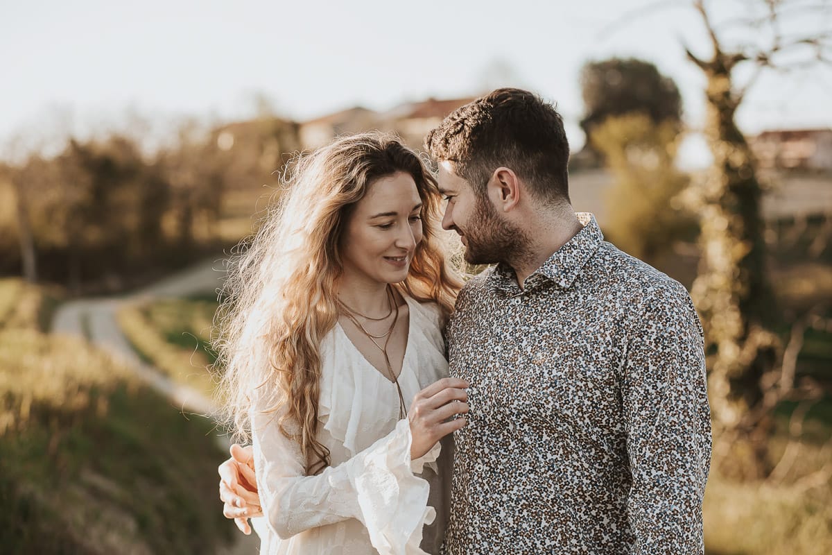 Anita e Davide, futuri sposi, si tengono per mano davanti a una chiesetta romanica a Mombello di Torino, baciati dal sole primaverile del Monferrato.