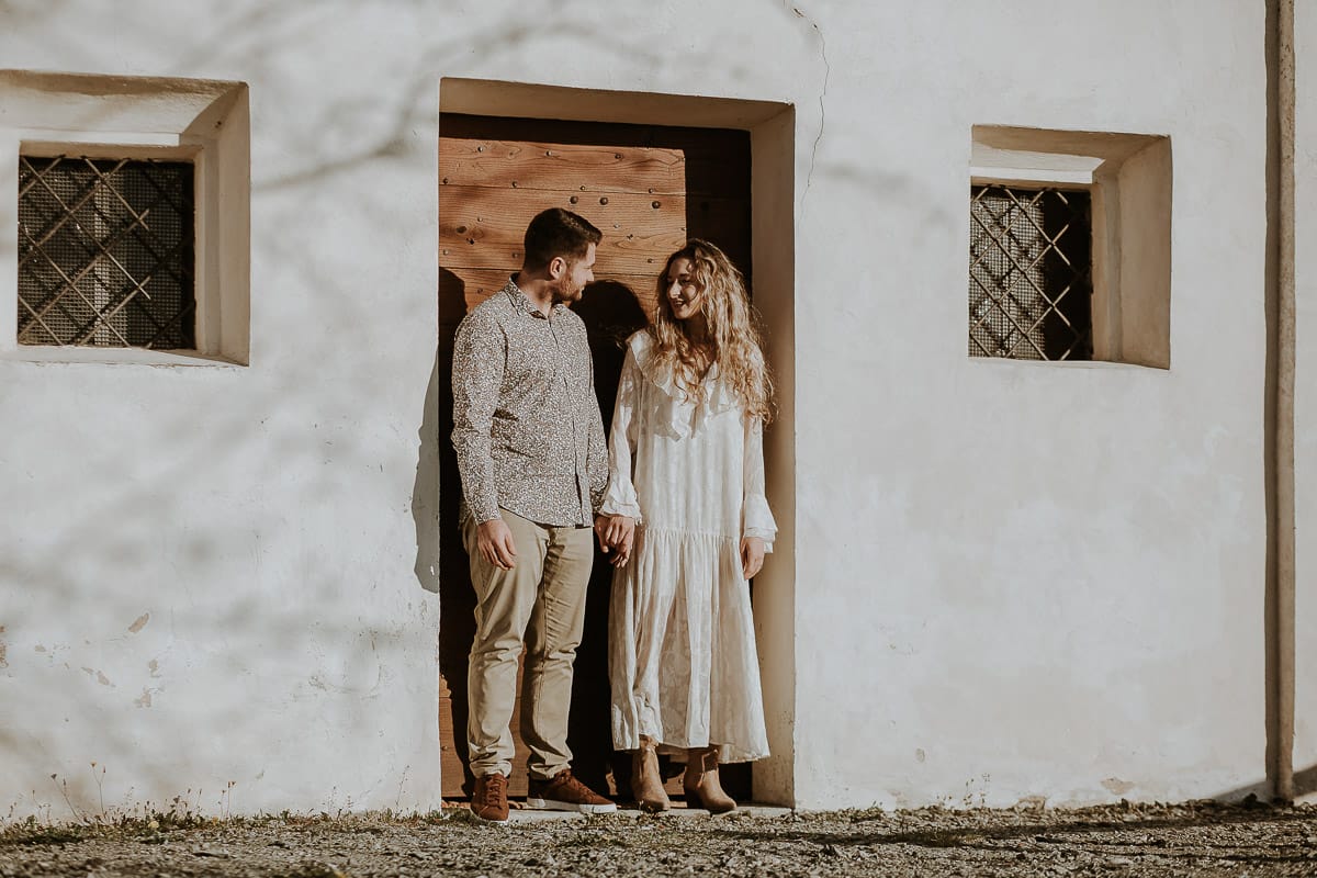Anita e Davide, futuri sposi, si tengono per mano davanti a una chiesetta romanica a Mombello di Torino, baciati dal sole primaverile del Monferrato.