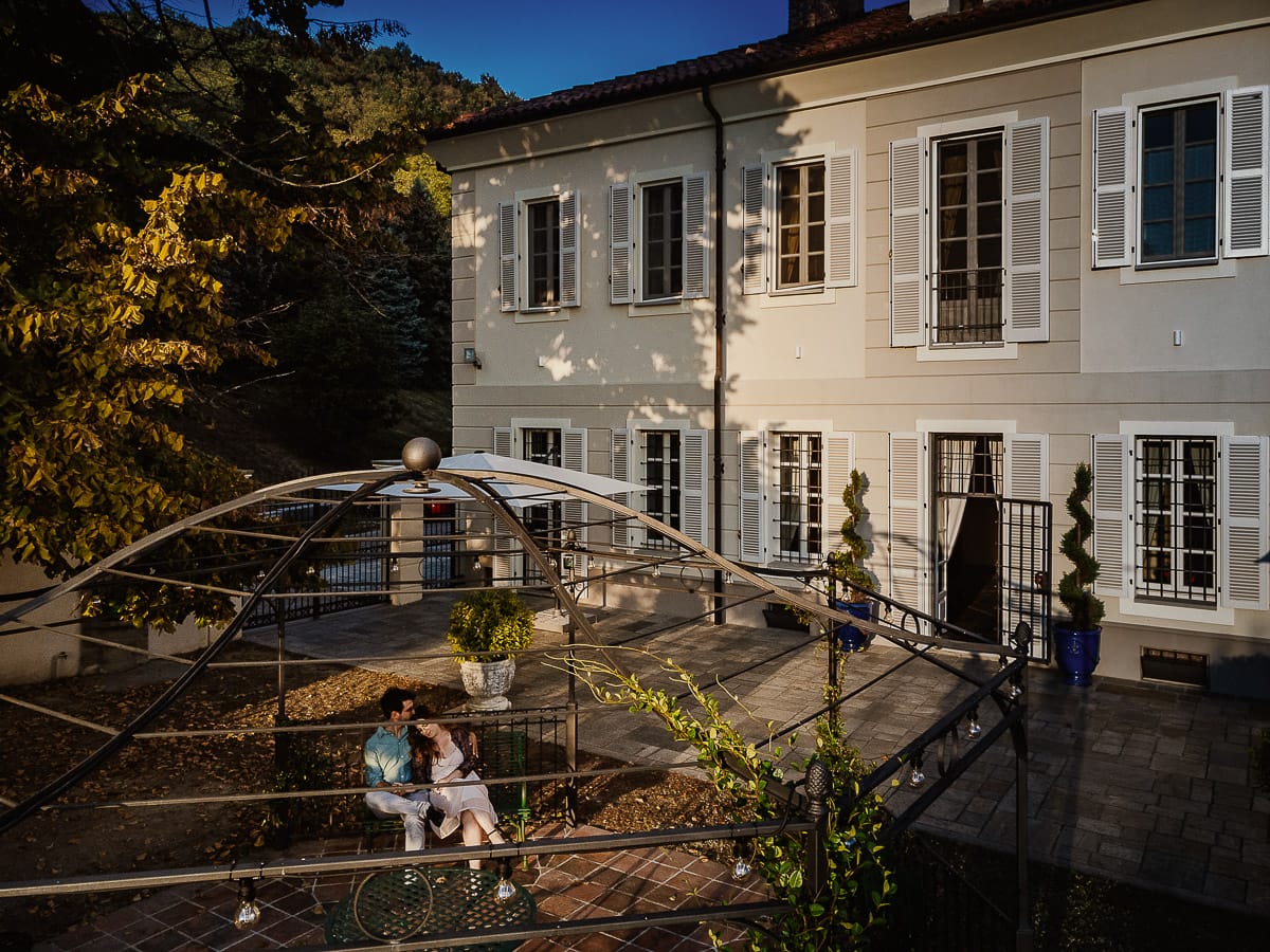 Fotografo matrimonio Rivalba. Villa Ala. Prewedding di Giorgia e Marco. Golden Hour. La coppia è seduti su di una panchina di ferro battuto, davanti alla villa sotto un gazebo in ferro battuto. Foto dal drone