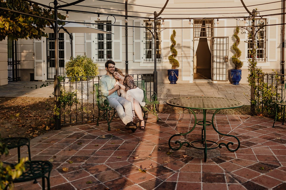 Fotografo matrimonio Rivalba. Villa Ala. Prewedding di Giorgia e Marco. Golden Hour. La coppia è seduti su di una panchina di ferro battuto, davanti alla villa sotto un gazebo in ferro battuto. Ritratto . Giorgia appoggia la testa sulla spalla di Marco.